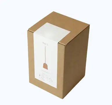 Medium-sized rectangular brown kraft corrugated cardboard box with white label wrapped around the top showing 'Koji S' text and a minimalist thin-line illustration of a pendant lamp with cord